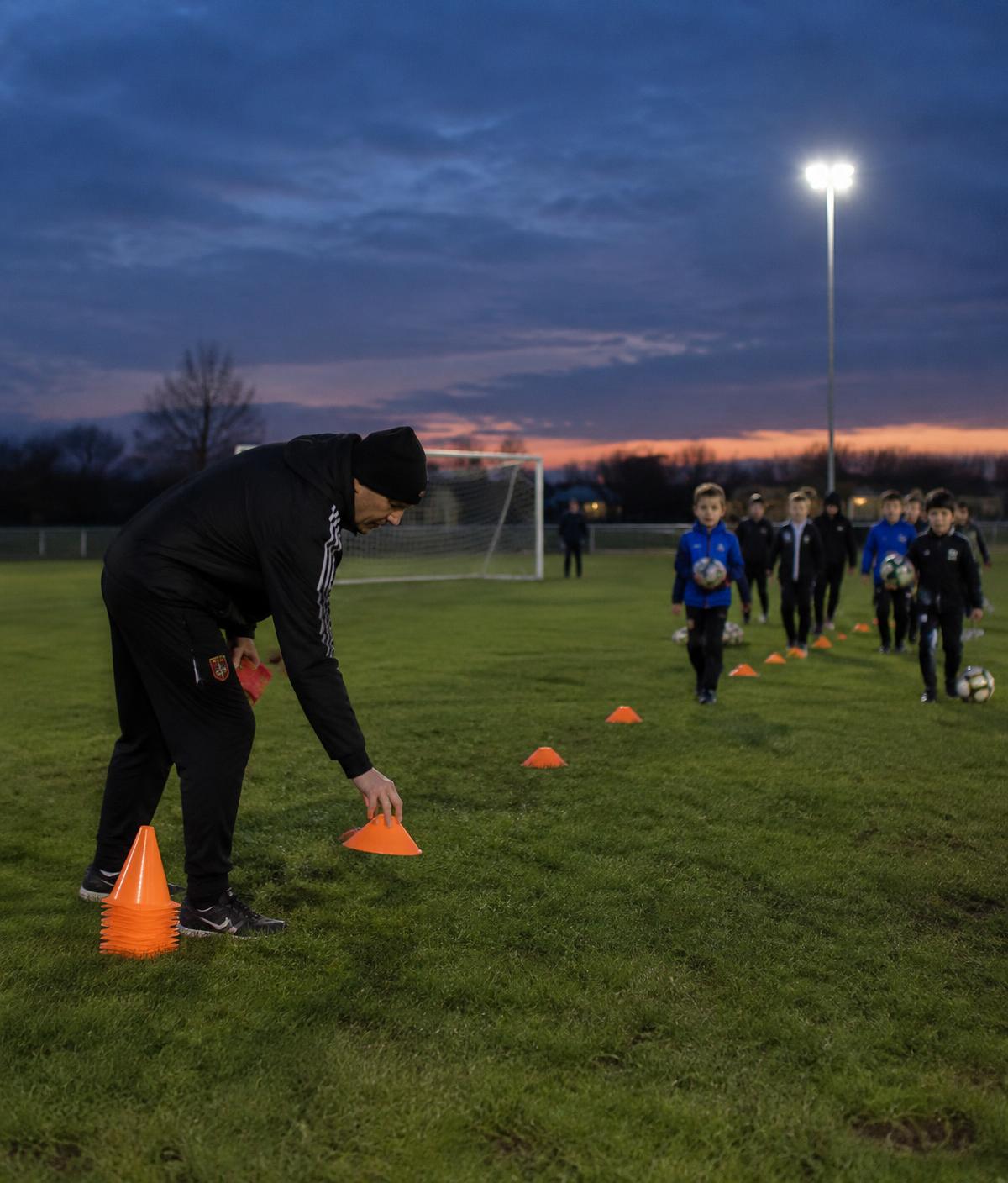 Coach setting out cones on a floodlit pitch at dusk